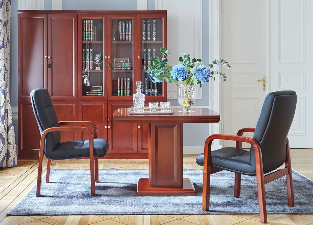 Elegancki stolik biurowy Visitor fornirowany drewnem w odcieniu koniaku. Stolik ma wymiary 100x100x75 cm. Kwadratowy blat na kolumnie zdobionej płycinami, rant frezowany. Klasyczny stolik do gabinetu.