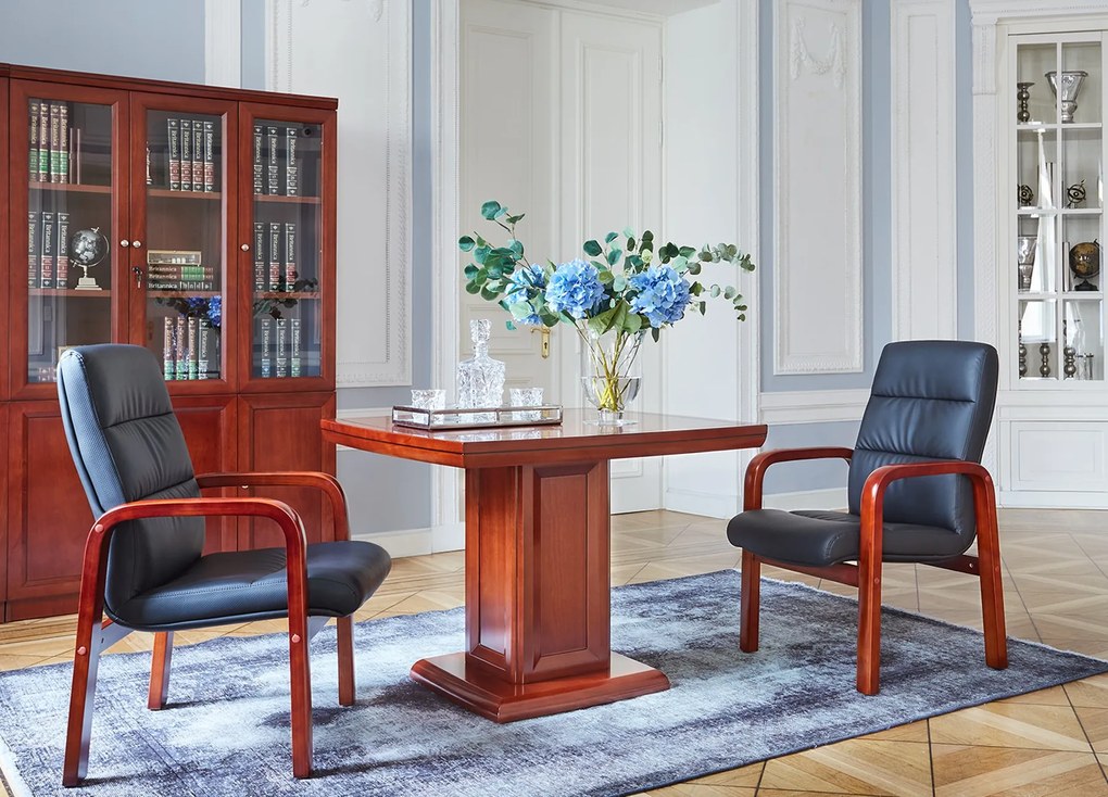 Elegancki stolik biurowy Visitor fornirowany drewnem w odcieniu koniaku. Stolik ma wymiary 100x100x75 cm. Kwadratowy blat na kolumnie zdobionej płycinami, rant frezowany. Klasyczny stolik do gabinetu.